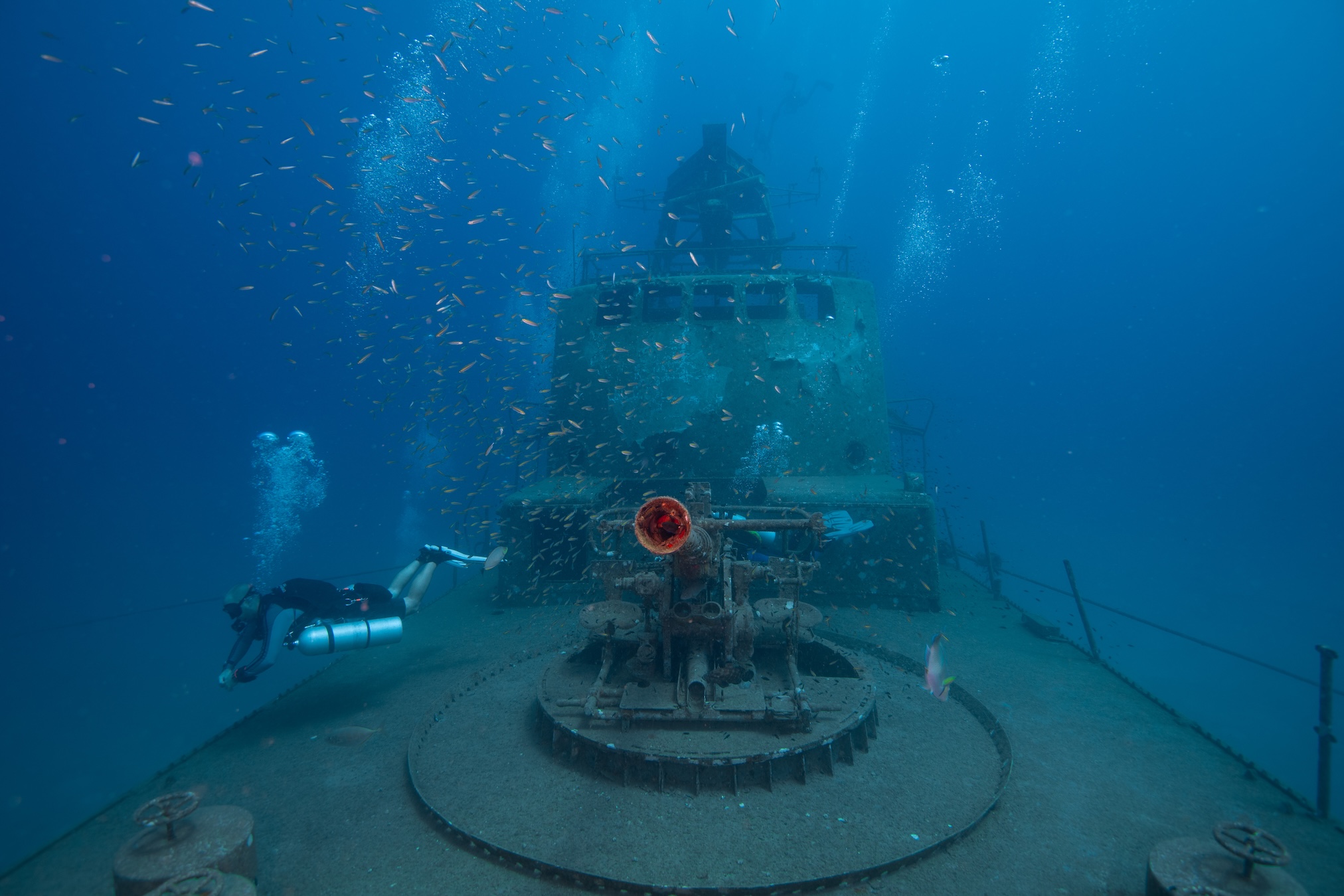 A scuba diver explores a sunken shipwreck near Koh Samui, surrounded by rust and vibrant marine life. Small fish dart around as the diver, clad in full gear, glides past the ship’s deck in the deep blue ocean waters, experiencing the wonders of Koh Samui diving.