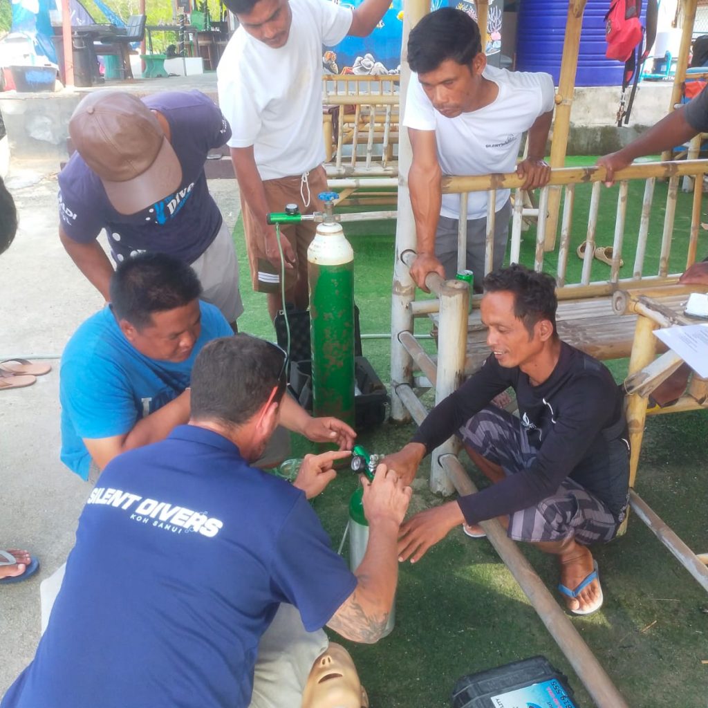 A group of men gather around a mannequin, practicing scuba diving safety procedures with an oxygen tank. They are in an outdoor setting, surrounded by bamboo structures and greenery, focused on the task at hand.