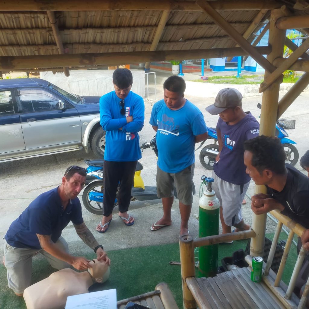 A group of people is gathered around a person demonstrating CPR techniques on a mannequin under a thatched roof structure. They are outside, with vehicles and scooters visible in the background. An oxygen tank is on the ground nearby.