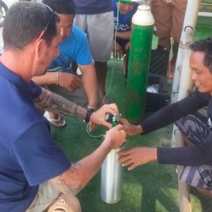 A group of people gather around to examine scuba diving equipment outdoors. A man kneels and works on a metal cylinder, while others watch intently. A diving mannequin lies on the ground nearby.