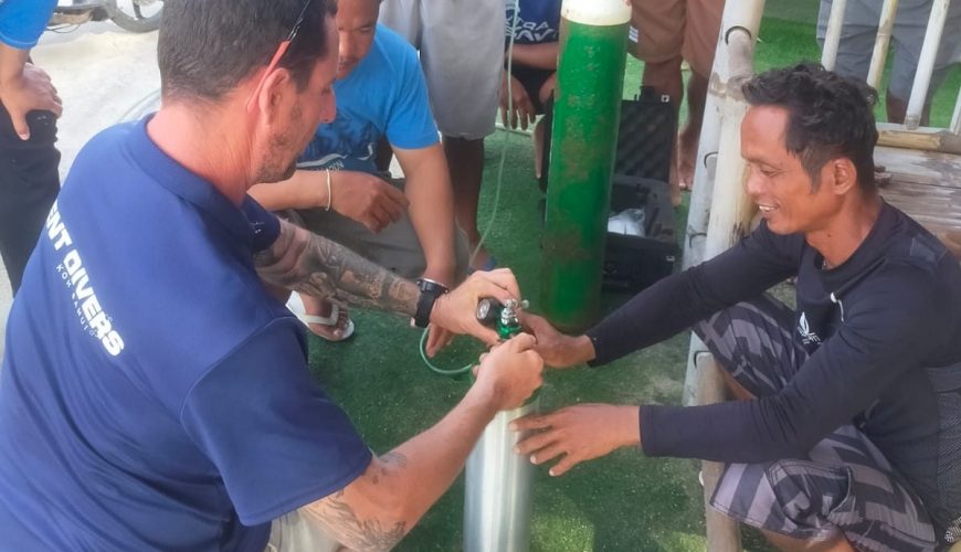 A group of people gather around to examine scuba diving equipment outdoors. A man kneels and works on a metal cylinder, while others watch intently. A diving mannequin lies on the ground nearby.