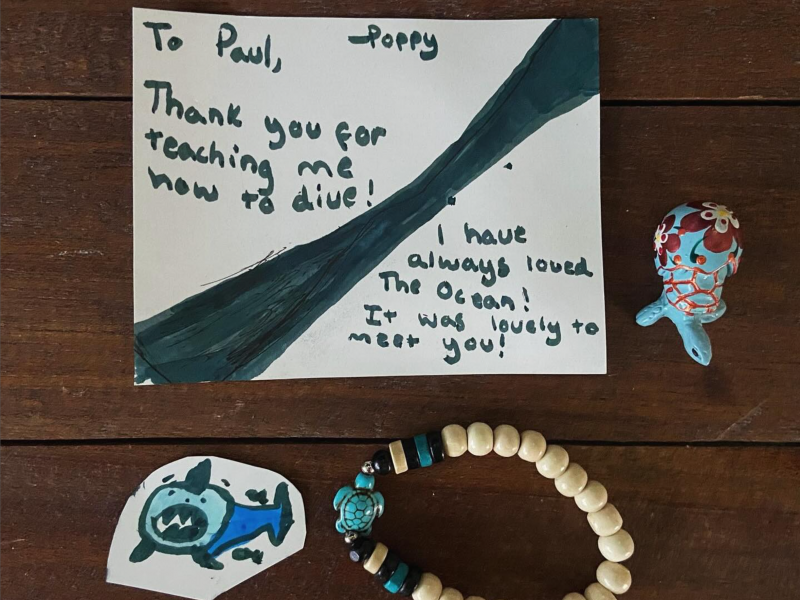 A handwritten thank you note addressed to Paul from Poppy, expressing gratitude for diving lessons. The note mentions love for the ocean. Nearby are a small colorful figurine, a shark drawing, and a bracelet with wooden beads on a wooden surface.