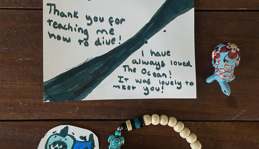 A handwritten thank you note addressed to Paul from Poppy, expressing gratitude for diving lessons. The note mentions love for the ocean. Nearby are a small colorful figurine, a shark drawing, and a bracelet with wooden beads on a wooden surface.