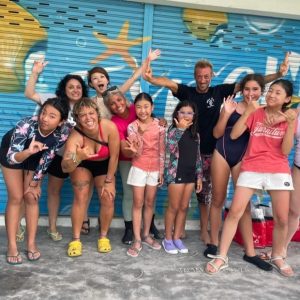 A group of people, including children and adults, pose happily in front of a colorful mural with fish. Some are waving, and there's a black dog on the left. The scene suggests a casual, joyful gathering.